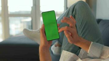 Man at home lying on a sofa and using smartphone with green mock-up screen in vertical mode. Guy browsing Internet video