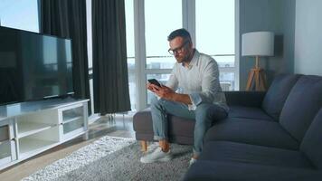Man with glasses sitting on the couch in a cozy room, speaking on a smartphone and using laptop. Remote work concept video