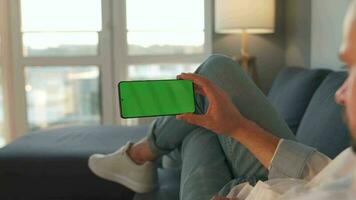 Man at home lying on a sofa and using smartphone with green mock-up screen in vertical mode. Guy browsing Internet video