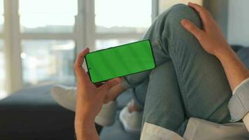 Man at home lying on a sofa and using smartphone with green mock-up screen in vertical mode. Guy browsing Internet video