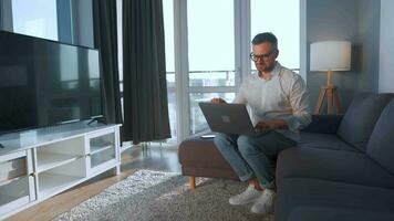 Man with glasses sitting on the couch in a cozy room and using smartphone for surfing internet or chatting in messengers video