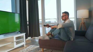 Man at home lying on a couch and watching TV with green mock-up screen video