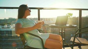 Woman drinks coffee and rests during a break on the balcony against the the setting sun. Remote work concept video