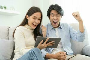Happy excited, smiling asian young couple love using tablet pc, great deal or business success, received or getting cash back, tax refund, good news by mail while sitting on sofa, couch at home. photo