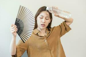 Summer heat stroke, hot weather, tired asian young woman sweaty and thirsty, refreshing with hand in blowing, wave fan to ventilation, holding cold water bottle tap her body when temperature high. photo