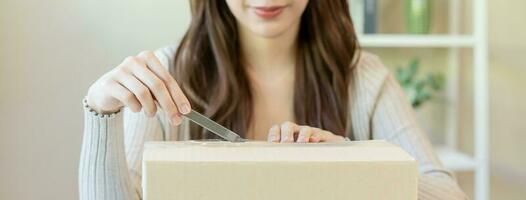 Happy asian young woman, girl customer sitting on sofa at home, hand in opening and unpacking cardboard box carton parcel after buying ordering present, shopping online, delivery service concept. photo