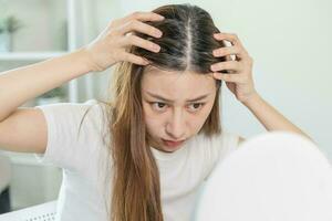 Damaged Hair, face serious asian young woman worry looking at scalp in mirror, hand in break into front hair loss, thin problem symptom at home. Health care shampoo beauty, isolated on background. photo