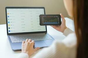 Investor analyzing exchange market, asian young business woman trader on graphic charts on laptop computer, hand in using smartphone for trading stock, looking at screen with diagrams at home office. photo