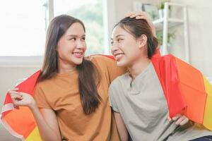 Happy lesbian, beautiful asian young two women, girl gay, couple love moment spending good time together, holding or waving lgbt rainbow, pride flag together, sitting on sofa at home. LGBT concept. photo