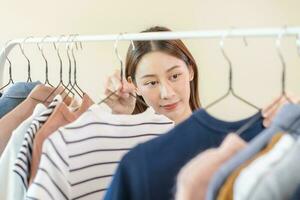Choice of clothes,Nothing to wear. Attractive asian young woman, girl try on appare, happy choosing dress, outfit on hanger in wardrobe in room closet at home. Deciding blouse what to put on which one photo