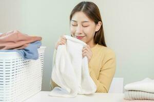 Feel softness, smiling asian young woman, girl touching fluffy towel cotton, smelling fresh clean clothes on table after washing, laundry, dry. Household working at home. Laundry and maid. photo