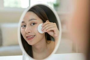 Happy beauty, beautiful asian young woman, girl smiling, looking in mirror, holding cotton pad, applying lotion by wipe on her face, removing makeup before shower at home. Skin care routine people. photo