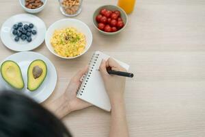 dieta, dieta mano de asiático joven mujer, niña escribir dieta plan nutrición en mesa es diferente comida ingredientes en el verde. nutricionista de saludable, nutrición de peso pérdida, salud cuidado gente. foto