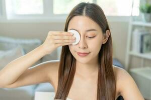 contento belleza, hermosa asiático joven mujer, niña mirando en a espejo, participación algodón almohadilla, aplicando facial limpiar en su rostro, quitando maquillaje antes de ducha en baño, piel cuidado en blanco antecedentes. foto