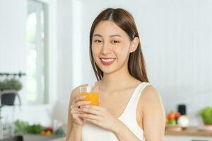 contento ama de casa, sonrisa asiático joven mujer, niña Bebiendo Fresco naranja jugo en un vaso bebida en el cocina, participación transparente vaso en su mano, sediento a hogar. salud cuidado, sano dieta concepto. foto