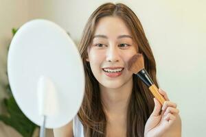 Happy routine beauty concept, pretty asian young woman, girl make up face by applying powder foundation by brush around face, looking at the mirror at home. Female look with natural fashion style. photo