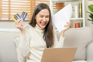 Happy excited, asian young woman, girl holding credit card and paperwork getting, received job promotion, approve tax refund cash back, looking good news at laptop computer, sitting on sofa at home. photo