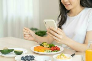 Dieting, asian young woman eating, holding fork at broccoli, diet plan nutrition with fresh vegetables salad, enjoy meal while using smartphone. Nutritionist of healthy, nutrition of weight loss. photo