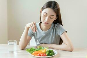 dieta en aburrido rostro, infeliz hermosa asiático joven mujer, niña en haciendo dieta, participación tenedor en ensalada lámina, disgusto o cansado con comer Fresco vegetales. nutrición de limpio, sano comida bueno gusto. foto