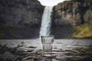 A glass sitting in front of a waterfall, photo