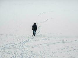 A man in a black jacket with a backpack walking on snow, footprints in snow, behind photo