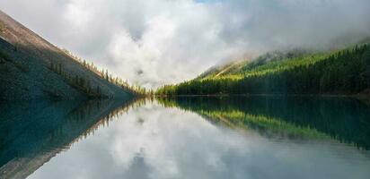 Morning panoramic landscape with dense fog over the lake. Reflection of coniferous trees in shiny calm water. Alpine tranquil landscape at early morning. Ghostly atmospheric scenery. photo