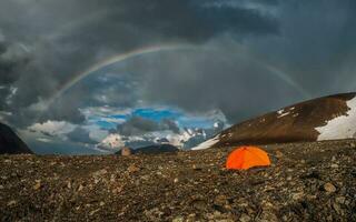 Panorama with a rainbow and an orange tent in the mountains. Atmospheric alpine landscape with snowy mountains with rainbow in rainy and sunny weather. photo