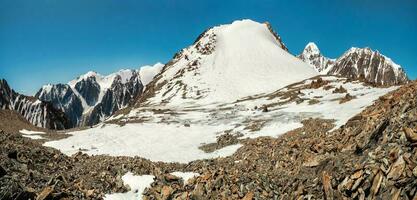 Snowy high-altitude plateau. Panoramic alpine landscape with snow-capped mountain peak and sharp rocks under blue sky. Colorful sunny mountain scenery with snow mountain top. photo