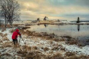 Photographer shoots a wonderful dawn snowy winter landscape with authentic house on the shore. photo