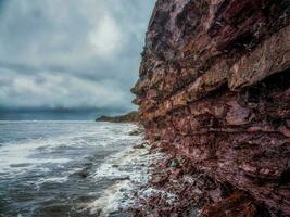 A cliff above the sea with a narrow coastline. Waves with white foam roll on the rocky shore. photo