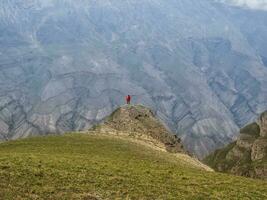 Small silhouette of woman with a backpack stands with her back over the cliff. Solitude in the mountains, solo travel concept. photo