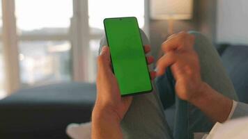 Man at home lying on a sofa and using smartphone with green mock-up screen in vertical mode. Guy browsing Internet video