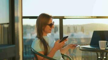 Frau Sitzung auf das Balkon gegen das Hintergrund von das Rahmen Sonne und macht ein online Kauf mit ein Anerkennung Karte video