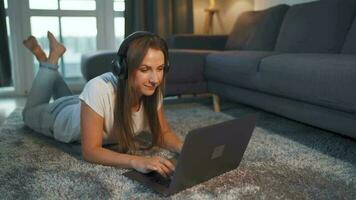 Woman is lying on the floor, working on a laptop and listens to music on headphones. Concept of remote work. video