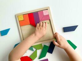 childs hand collects multicolored wooden mosaic on white background. child solves colorful tangram photo
