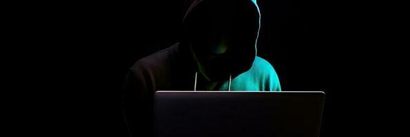 Hacker without face in a hood holds a laptop. on a dark background. Banner for cyber security.  Internet web hack technology. Digital laptop in hacker man hand isolated on black. photo
