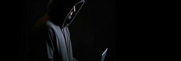 Hacker without face in a hood holds a laptop. on a dark background. Banner for cyber security.  Internet web hack technology. Digital laptop in hacker man hand isolated on black. photo