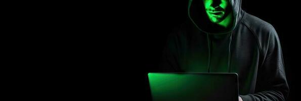 Hacker without face in a hood holds a laptop. on a dark background. Banner for cyber security.  Internet web hack technology. Digital laptop in hacker man hand isolated on black. photo