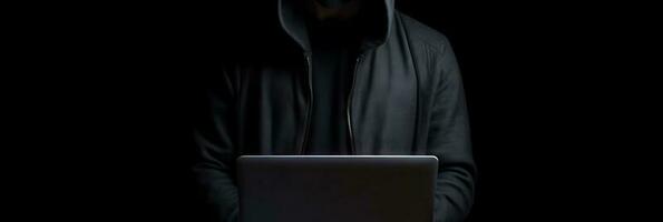 Hacker without face in a hood holds a laptop. on a dark background. Banner for cyber security.  Internet web hack technology. Digital laptop in hacker man hand isolated on black. photo