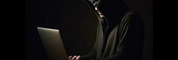 Hacker without face in a hood holds a laptop. on a dark background. Banner for cyber security.  Internet web hack technology. Digital laptop in hacker man hand isolated on black. photo