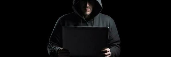 Hacker without face in a hood holds a laptop. on a dark background. Banner for cyber security.  Internet web hack technology. Digital laptop in hacker man hand isolated on black. photo
