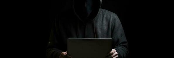 Hacker without face in a hood holds a laptop. on a dark background. Banner for cyber security.  Internet web hack technology. Digital laptop in hacker man hand isolated on black. photo