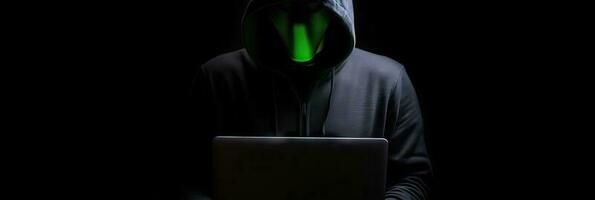 Hacker without face in a hood holds a laptop. on a dark background. Banner for cyber security.  Internet web hack technology. Digital laptop in hacker man hand isolated on black. photo