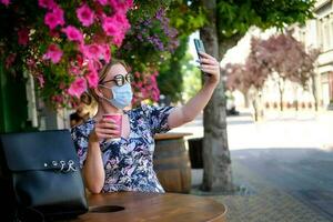 Beautiful young woman in a cafe in a sunglasses and medical mask and makes a selfie on the phone. A girl in a cafe on the street communicates online on a smartphone. Pandemic covid-19, quarantine. photo