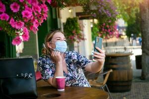 Beautiful young woman in a cafe in a medical mask and makes a selfie on the phone. A girl in a cafe on the street communicates online on a smartphone. Pandemic covid-19, quarantine. photo