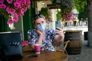 Beautiful young woman in a cafe in a medical mask and makes a selfie on the phone. A girl in a cafe on the street communicates online on a smartphone. Pandemic covid-19, quarantine. photo