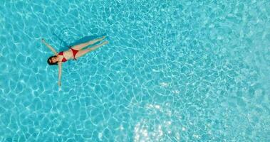 topo baixa Visão do uma mulher dentro vermelho roupa de banho deitado em dela costas dentro a piscina. video