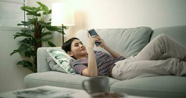 joven asiático hembra sonriente y surf el Internet en un móvil teléfono mientras acostado en el sofá foto