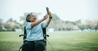maduro asiático mujer en silla de ruedas utilizar teléfono inteligente hacer vídeo llamada foto