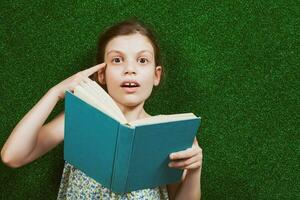 pequeño niña es acostado en artificial césped con un libro foto